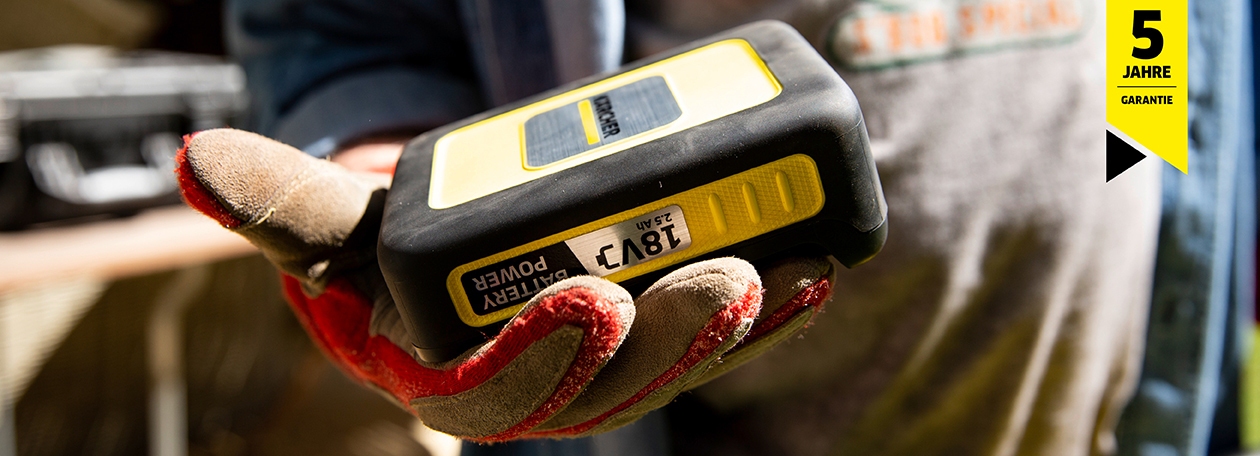 Der Kärcher 18V Battery Power Akku: Power für jedes Projekt Eine Person mit Arbeitshandschuhen hält einen gelb-schwarzen Kärcher 18V Battery Power Wechselakku mit 2,5 Ah Kapazität. Im Hintergrund ist ein gelbes Siegel mit der Aufschrift „5 Jahre Garantie“ zu sehen.
