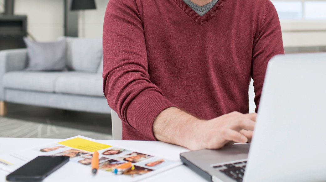Eine Person in einem weinroten Pullover sitzt an einem weißen Schreibtisch und bedient einen Laptop, um online Mietgeräte der Alfred Kärcher GmbH zu reservieren.