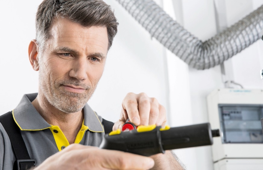Nahaufnahme eines Servicetechnikers der Alfred Kärcher GmbH in grau-gelber Arbeitskleidung, der konzentriert ein schwarzes Bauteil eines Reinigungsgeräts prüft.