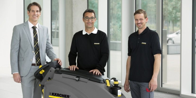 Drei kompetente Kärcher Experten stehen hinter einer großen, grau-schwarzen Scheuersaugmaschine in einem hellen, modernen Gebäude.