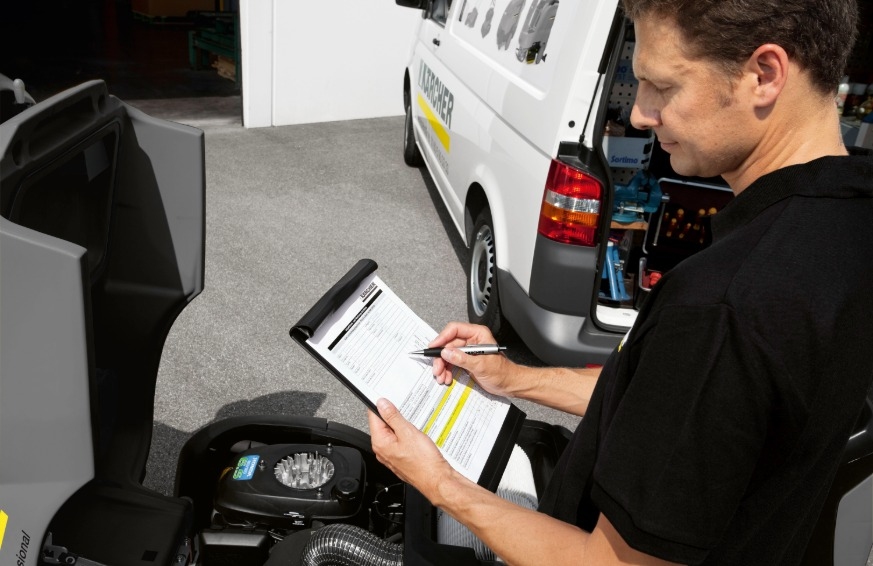 Ein Servicetechniker der Alfred Kärcher GmbH in schwarzer Arbeitskleidung füllt konzentriert eine Inspektions-Checkliste auf einem Klemmbrett aus. Er steht im Freien neben einer Reinigungsmaschine mit geöffneter Wartungshaube, während im Hintergrund ein w