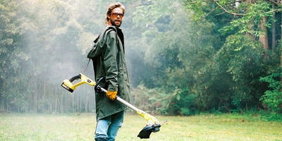Leistungsstarke Kärcher Gartengeräte für die anspruchsvolle Grundstückspflege Ein Mann in einem grünen Parka, Schutzbrille und Handschuhen steht in einer nebligen Wald- oder Gartenumgebung und hält einen gelb-schwarzen Akku-Rasentrimmer von Kärcher.