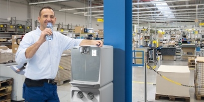 Robuste Kärcher Wasserversorgung Ein Mitarbeiter in Arbeitskleidung trinkt in einer großen Produktionshalle Wasser aus einer Flasche neben einem stationären Kärcher Wasserspender.