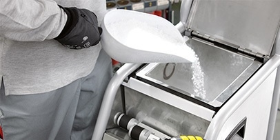 Befüllen eines Trockeneisstrahlgeräts mit Pellets Eine Person in grauer Arbeitskleidung und schwarzen Schutzhandschuhen gießt mit einer großen weißen Schaufel Trockeneis-Pellets in den geöffneten Behälter eines professionellen Reinigungsgeräts.