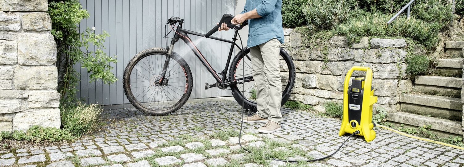 Een man reinigt zijn fiets met een draadloze gele Kärcher hogedrukreiniger op een oprit van kasseien