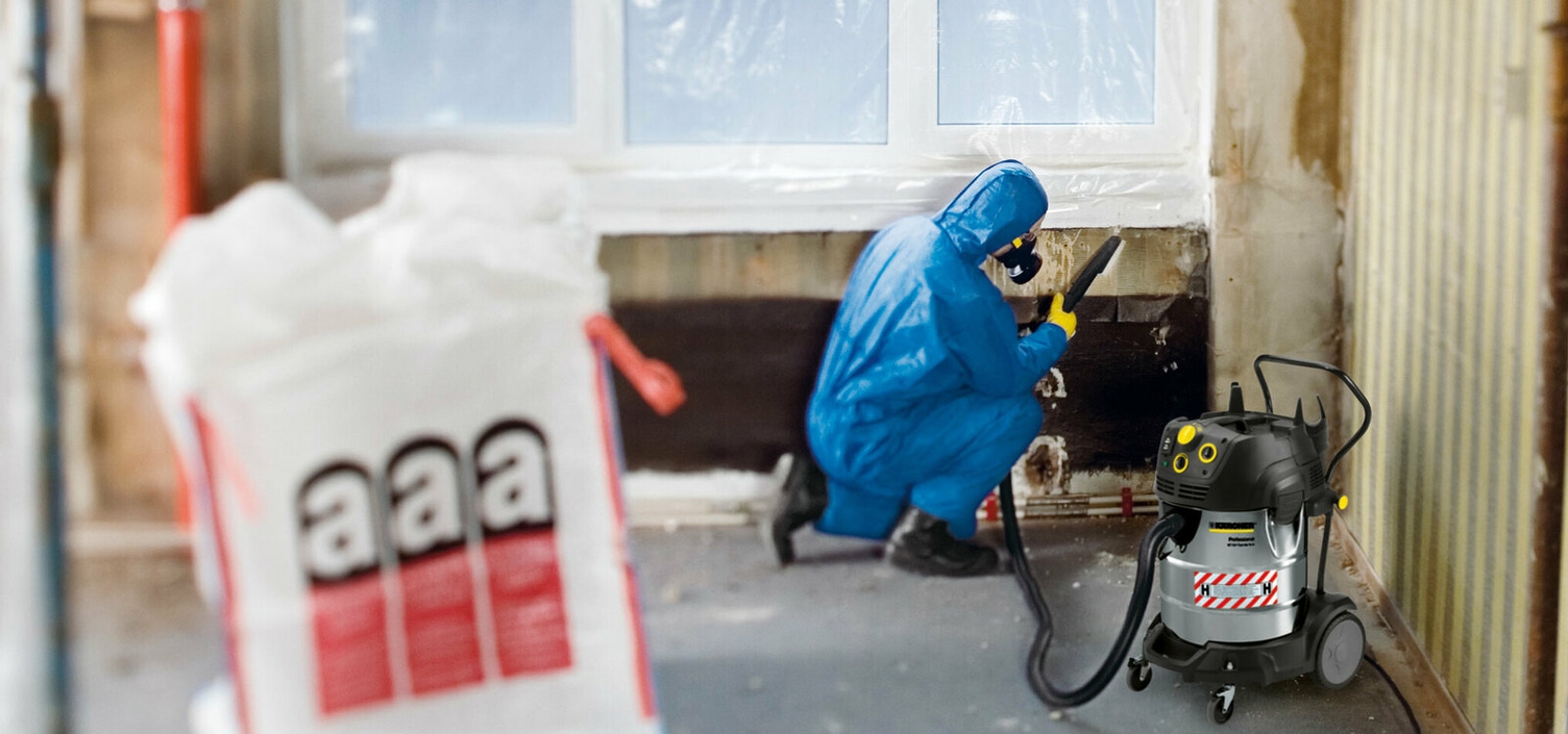 Asbest afzuigen Een persoon in blauwe veiligheidskleding met ademhalingsmasker zuigt vuilresten van een ruwbouwmuur op met een Kärcher veiligheidsstofzuiger