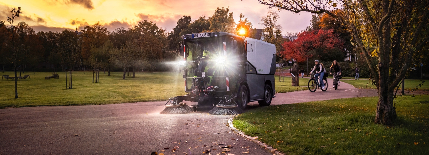 Elektrische MC 250 Een elektrische Kärcher straatveegmachine met felle lichten reinigt een fietspad in een park tijdens zonsondergang