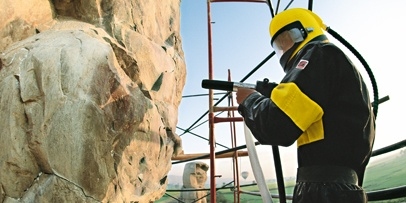 Nauwkeurige reiniging van het kwartsiet met gecontroleerde perslucht en ultrafijn calciumcarbonaat en Kärcher-technicus in een geel-zwart beschermend pak en helm reinigt het steenoppervlak van de Memnonkolossen met een straalpistool.