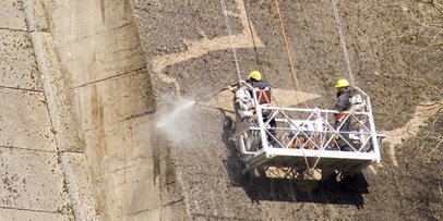 Twee technici spuiten vanuit een hangende werkgondel algen en mossen van de betonwand om diermotieven te creëren.