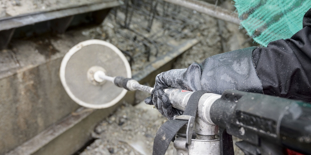 Een persoon in een beschermend pak houdt watergereedschap vast met een slang richting het beton]