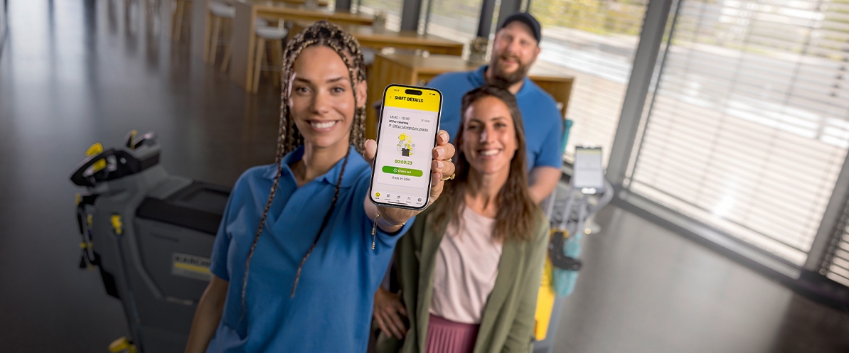 Lächelnde Reinigungskraft mit Scheuersaugmaschine nutzt Connected Cleaning auf ihrem Handy