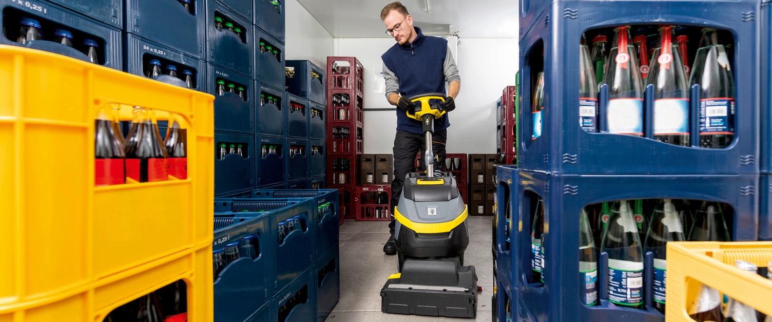 Kühlhaus reinigen mit Scheuersaugmaschine mit Ware Frontansicht eines Mannes, der mit einem Reinigungsgerät zwischen Getränkekisten sauber macht.