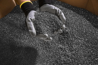 Mit Handschuhen bekleidete Hände, die eine Schaufel voll dunkles recyceltes Kunststoffgranulat halten, während sich darunter eine große Fläche des Materials ausbreitet