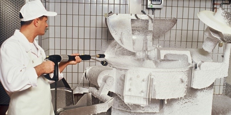 Ein Mann in weißer Arbeitskleidung sprüht mit einem Kärcher Hochdruckreiniger eine Metzgerei-Maschine ab