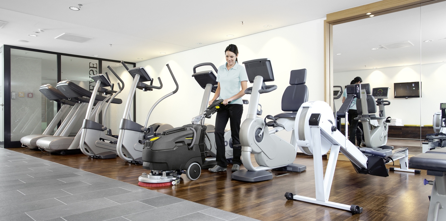 Scheuersaugmaschine Eine Frau reinigt mit einer Scheuersaugmaschine von Kärcher einen Fitnessbereich