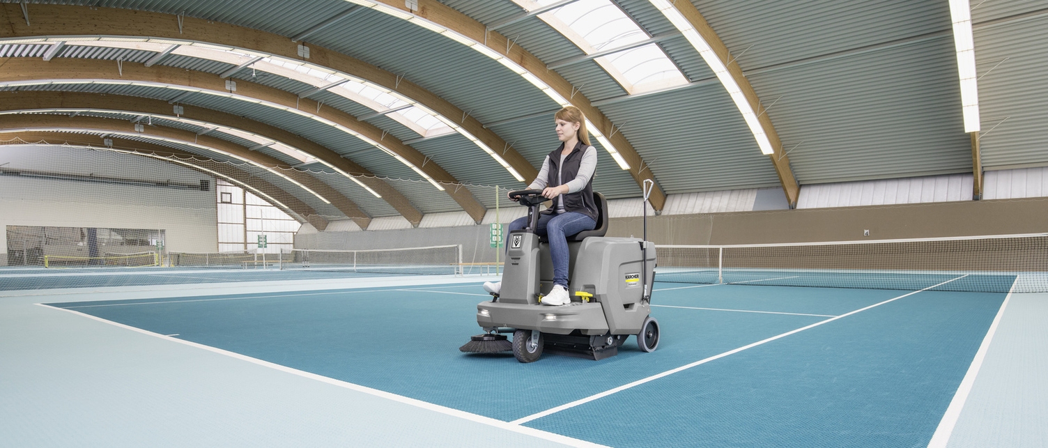 Teppichkehrsaugmaschine Eine Frau reinigt mit einer Aufsitz-Teppichkehrsaugmaschine den Boden eines Indoor-Tennisplatzes