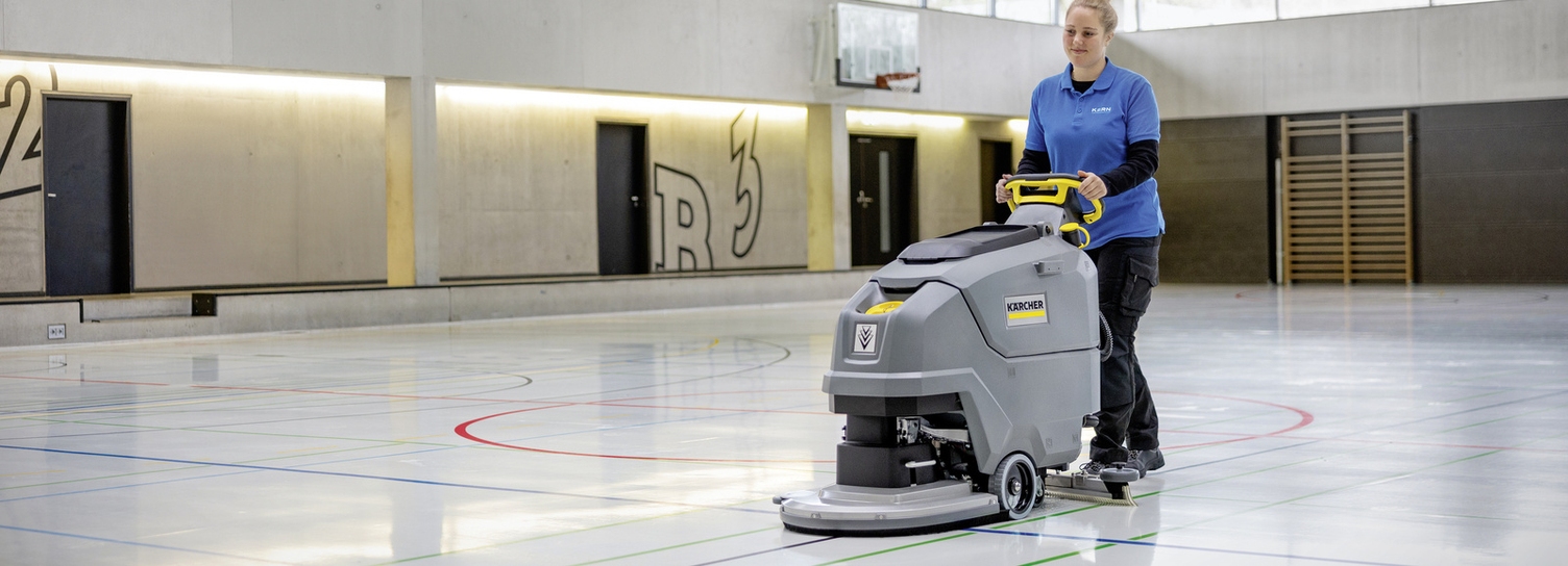 Scheuersaugmaschine im Einsatz Eine Frau reinigt mit einer großen Scheuersaugmaschine den Boden einer Turnhalle