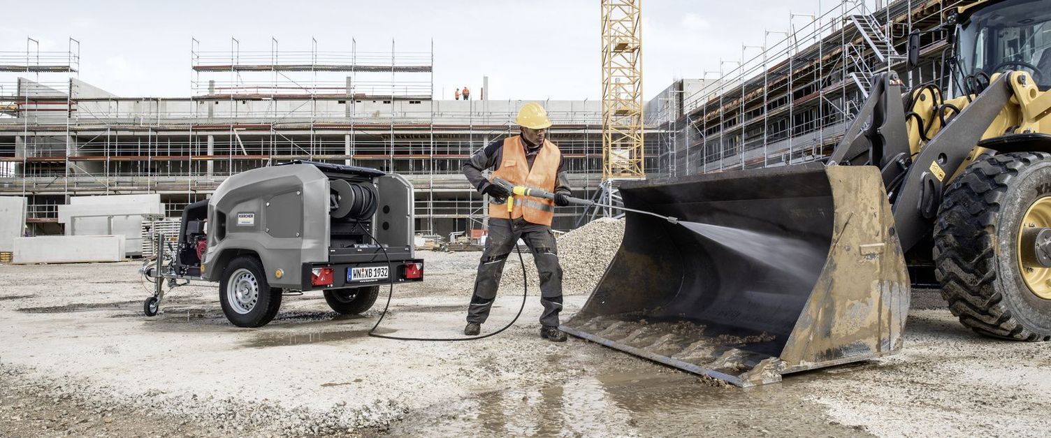 A person wearing a high-vis vest and a helmet cleans a construction machine using a high-pressure cleaner