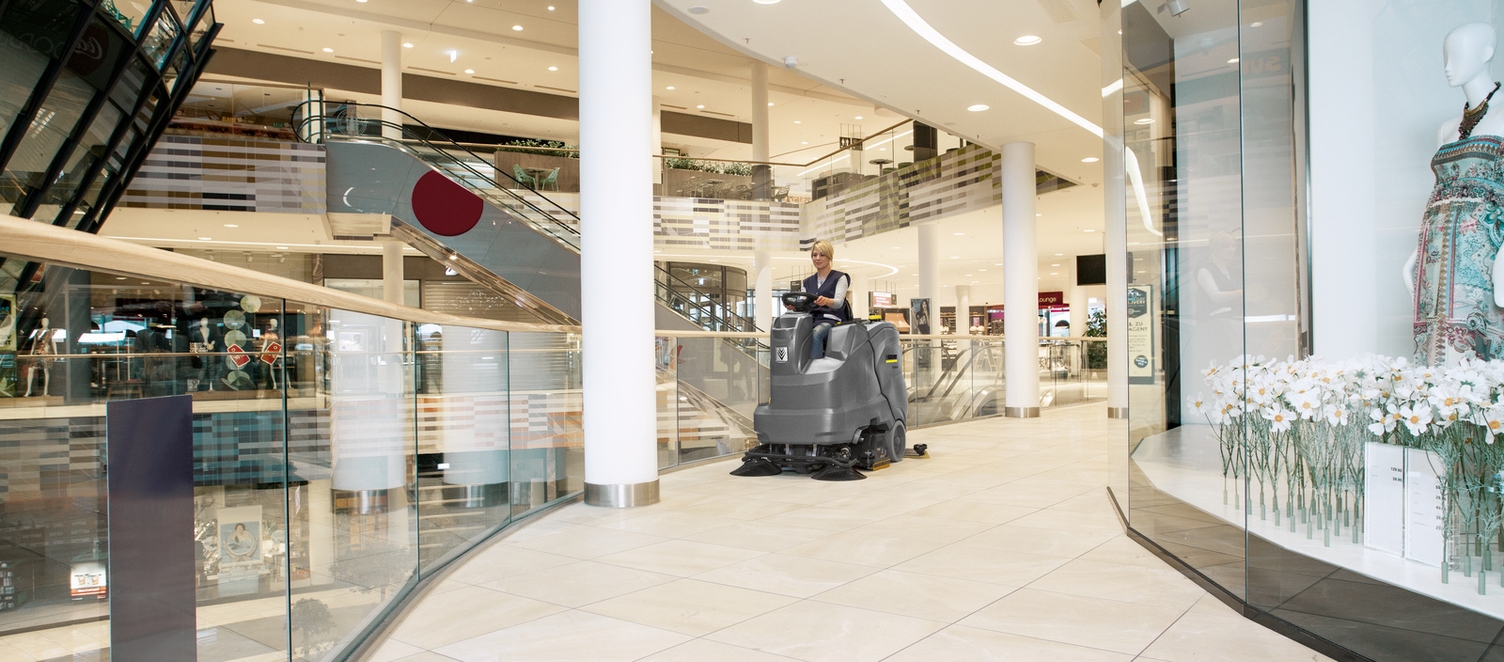 A person is cleaning a shopping area with a ride-on scrubber dryer