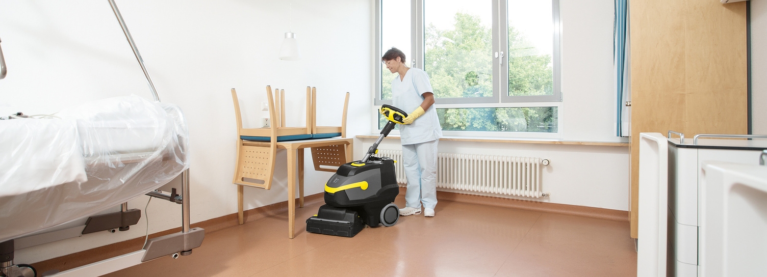 A woman cleaning the floor of a patient room with a scrubber dryer