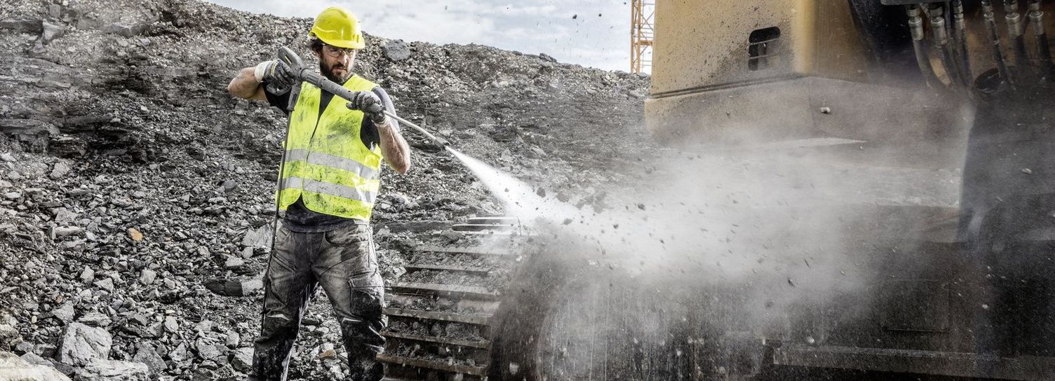 A person wearing a high-vis vest and a helmet uses a Kärcher High-Pressure Cleaner to clean a construction machine.
