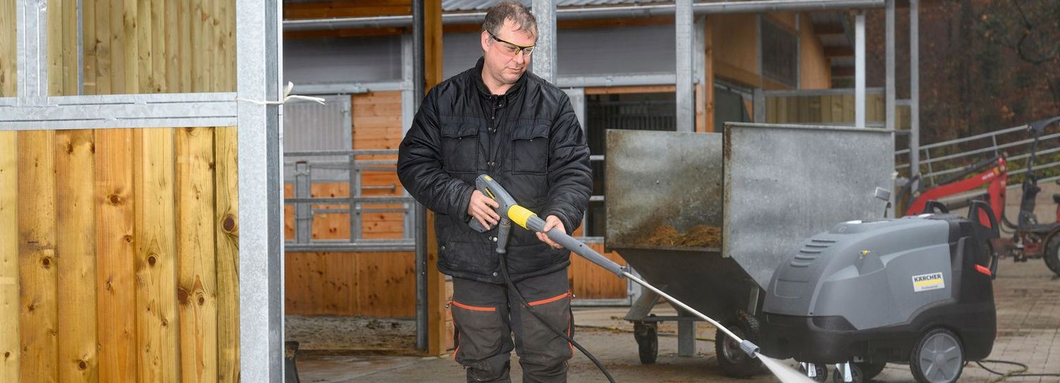 A man cleans the yard in front of the horse stable with a pressure washer