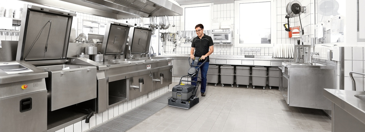 A person is cleaning the floor of a hospital kitchen using a scrubber dryer