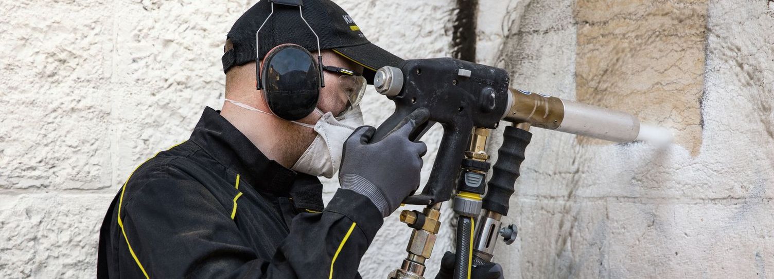 Abrasive particle blasting method for facade cleaning A person wearing earmuffs and gloves uses a particle blasting device to clean a facade