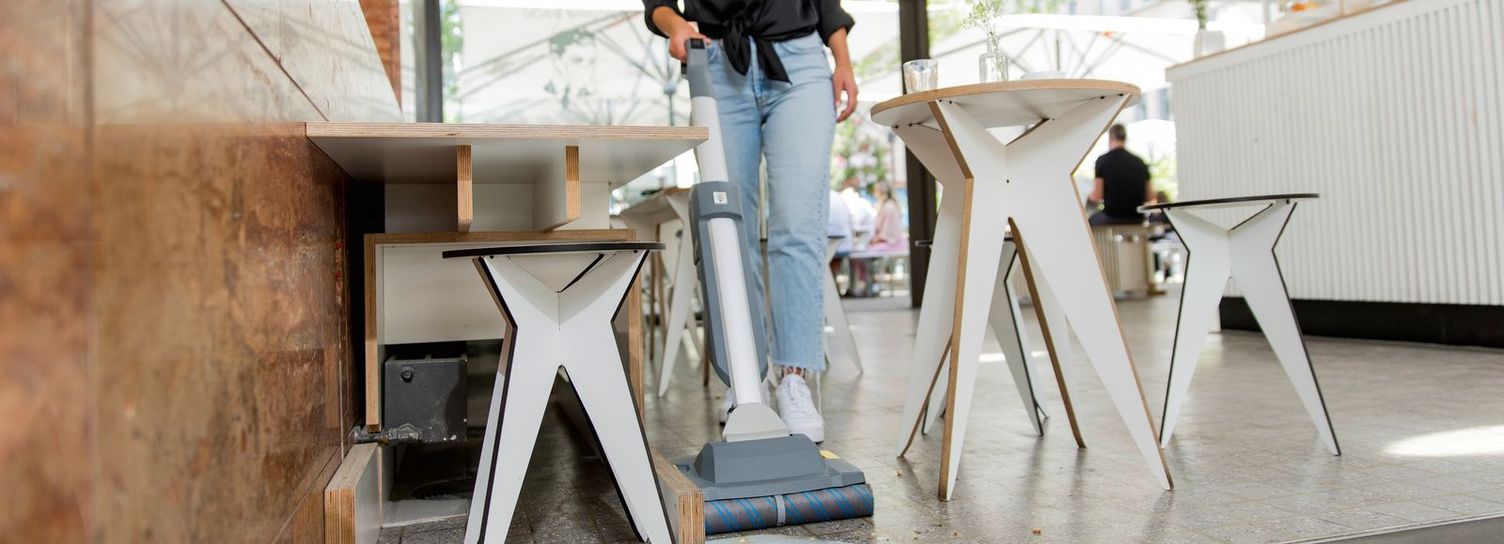 Café floor cleaning A woman using a vacuum cleaner to suck up a liquid between tables and chairs in a café.