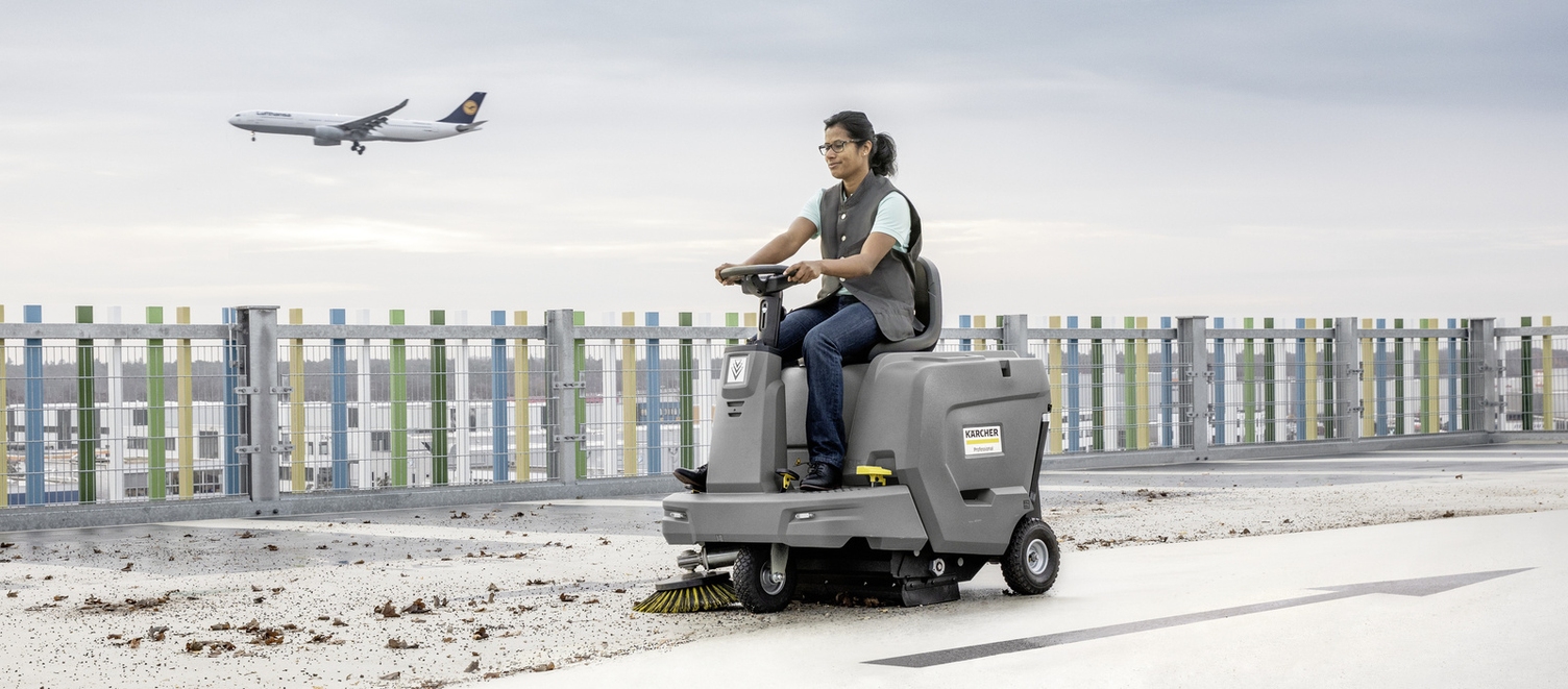 A person cleans the area around the airport using a ride-on sweeper