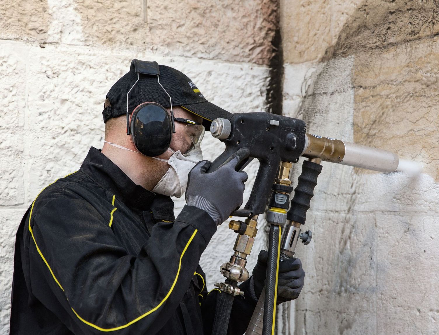 A person wearing protective eyewear, a mask and ear defenders cleans a natural stone wall using the low-pressure grit blasting cleaning method.