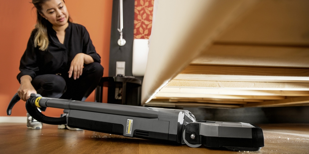 A person cleans the floor under a bed with the help of a carpet brush vacuum cleaner