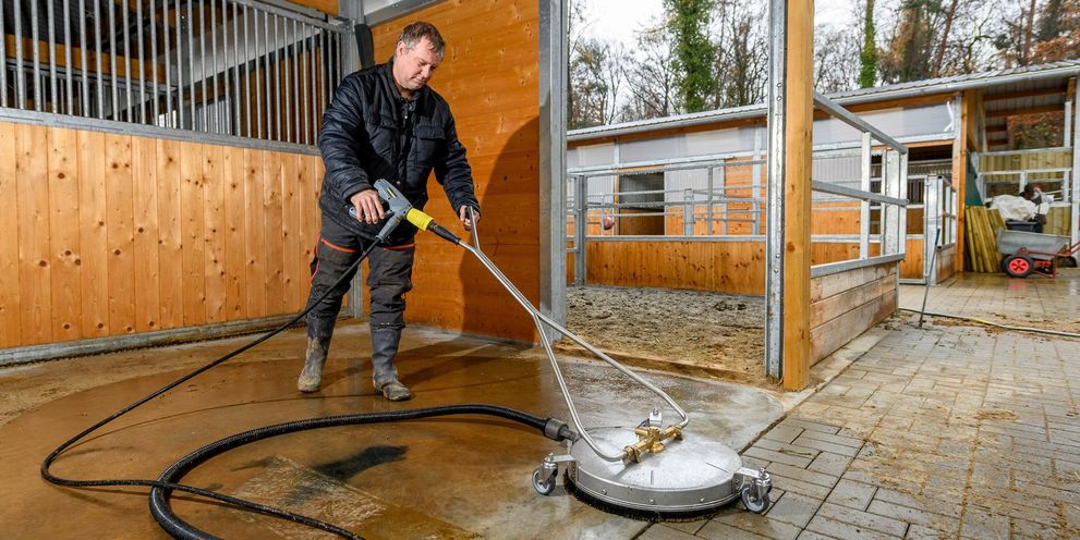 A man uses a Kärcher high-pressure cleaner with a surface cleaner attachment for wet cleaning in the horse stall.
