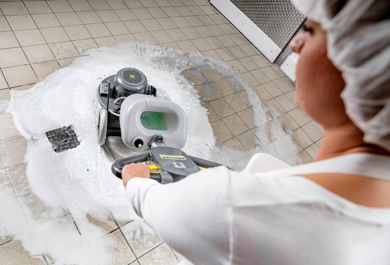 Floor cleaning in bakery View from above: A woman in white work clothes with a hood cleans the floor with foam using a Kärcher device