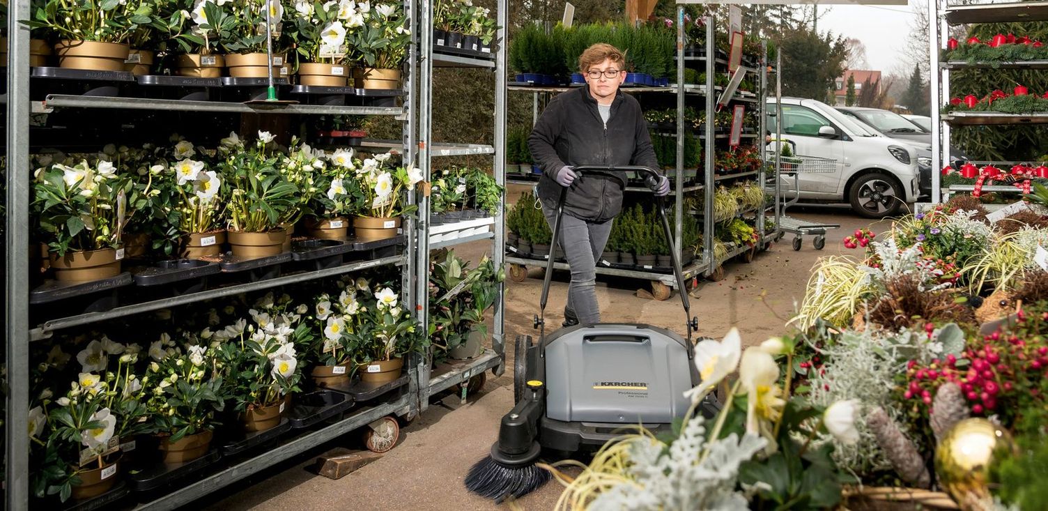 A woman cleans the floor between plant shelves with a Kärcher sweeper