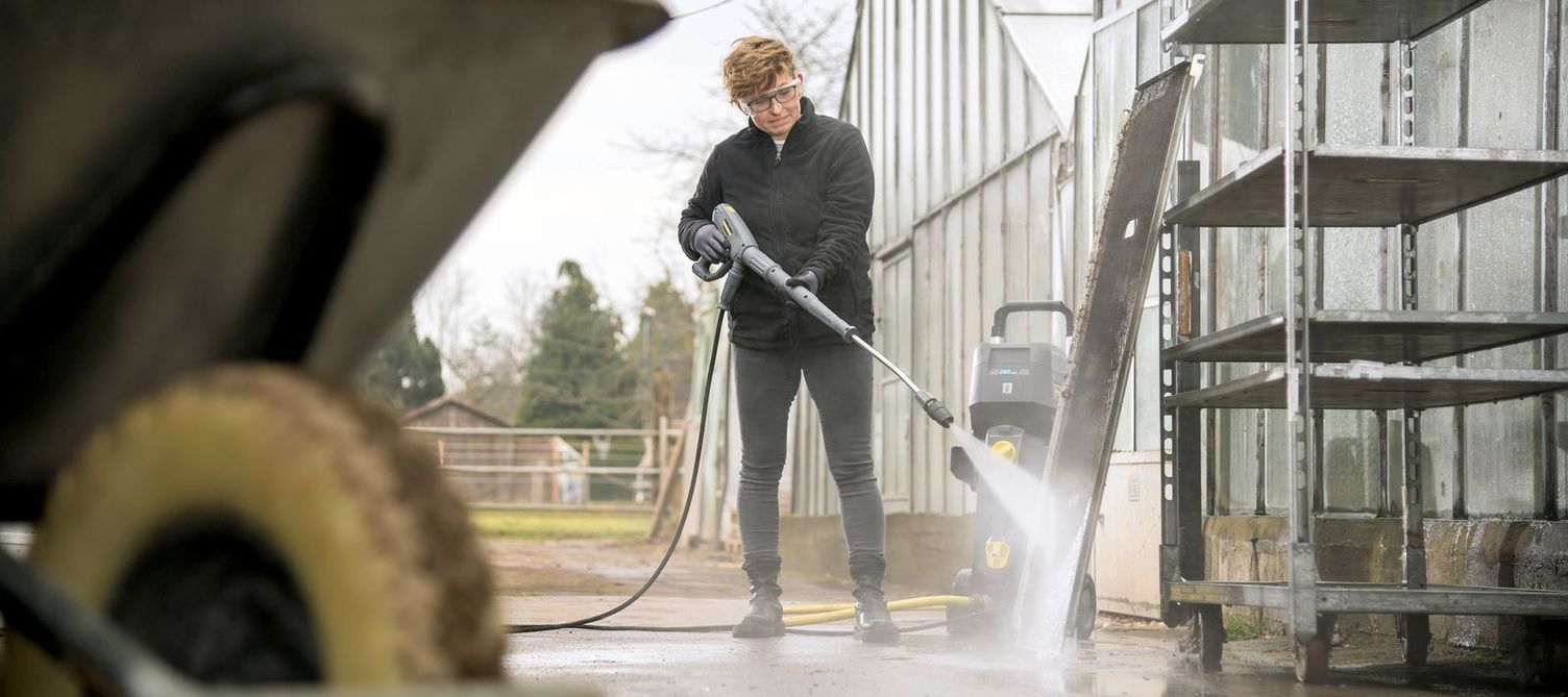 A man cleans outdoor shelves in front of a greenhouse with a Kärcher pressure cleaner