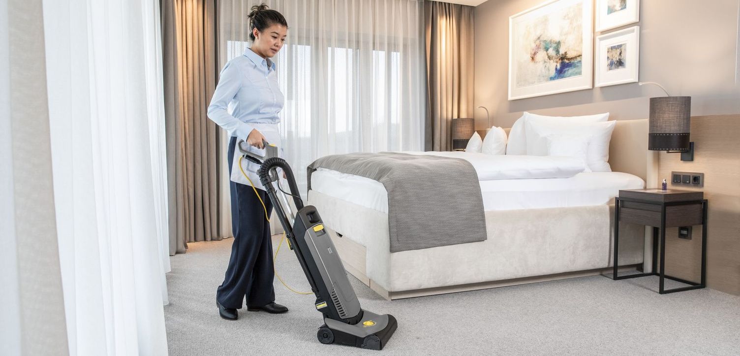 A service staff member cleans the carpet with a Kärcher vacuum cleaner in a hotel room.