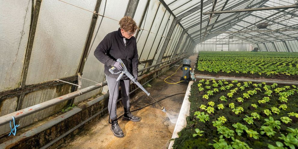 A man cleans dirt from a planting frame in a greenhouse with a Kärcher device.