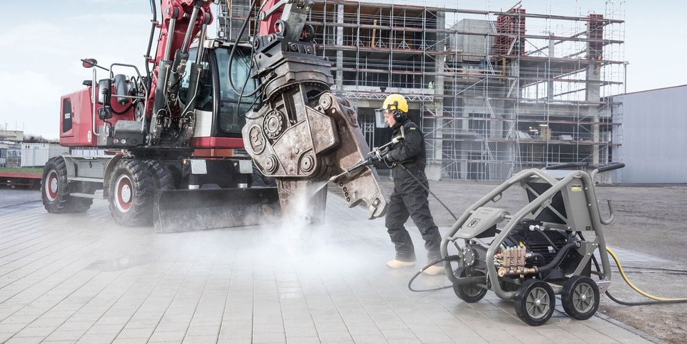 A person in protective clothing cleans a red construction machine with a Kärcher Ultra-High Pressure Cleaner