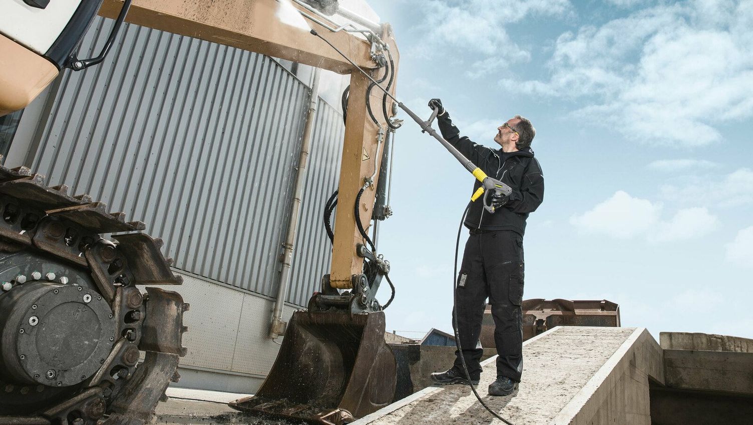 A person uses a Kärcher Hot Water High-Pressure Cleaner to wash an excavator