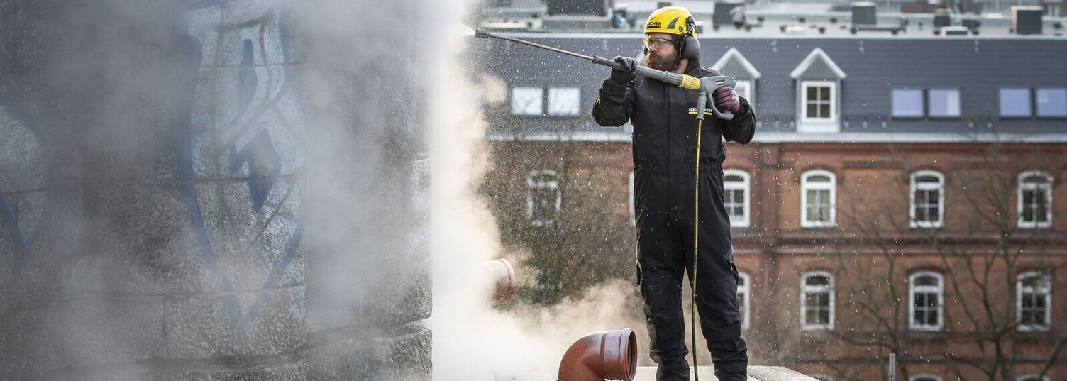 Pressure washing is one method for cleaning natural stone facades A person wearing protective clothing and a helmet is standing on a rooftop and cleaning a stone facade with a Kärcher Pressure Washer