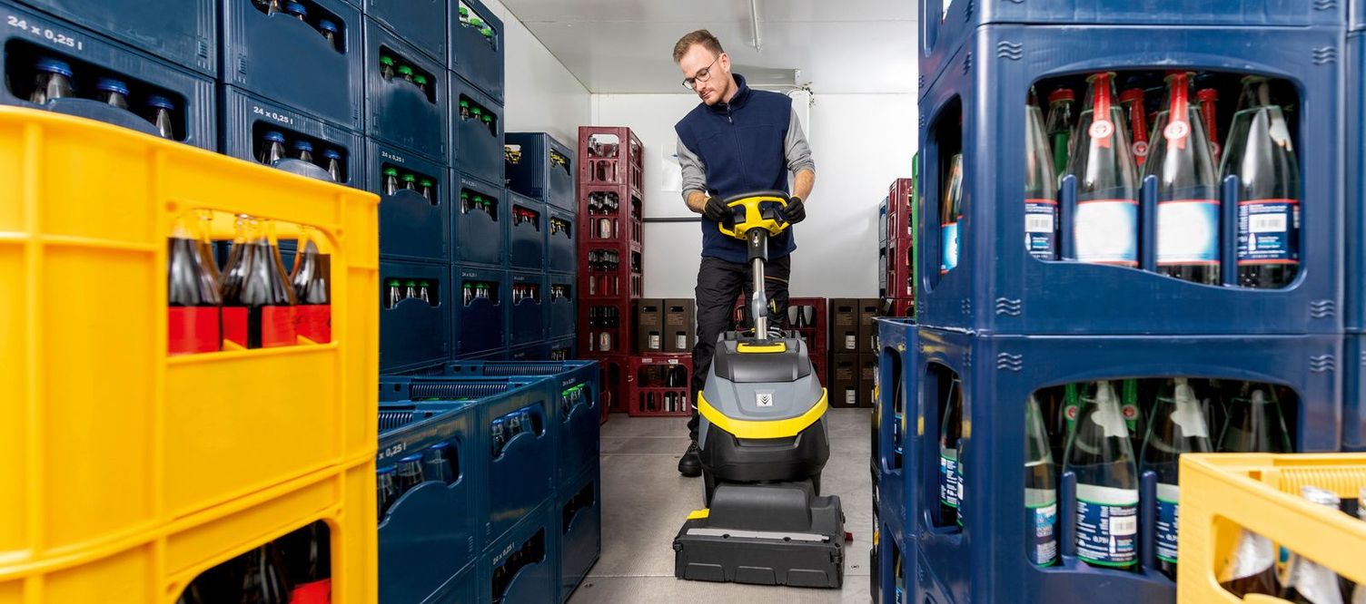 Front view of a man cleaning between crates of drinks with cleaning equipment.
