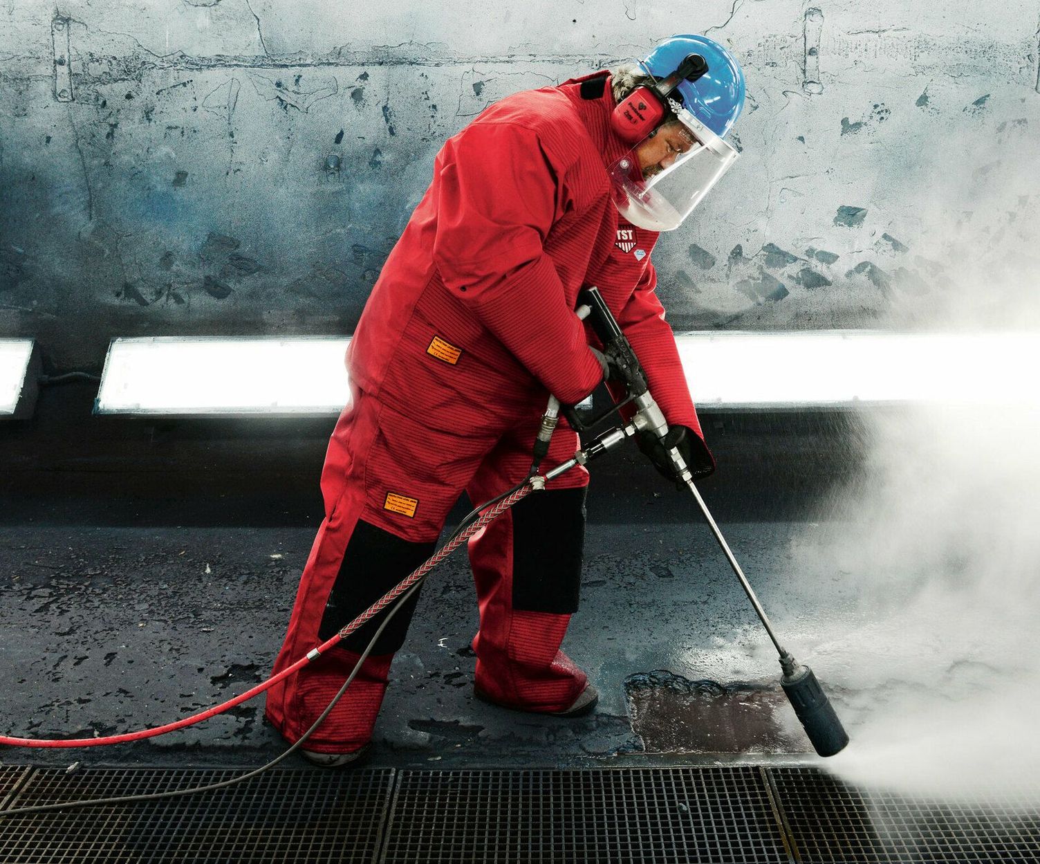 A person in a red protective suit uses ultra-high-pressure water jets on concrete in an industrial hall