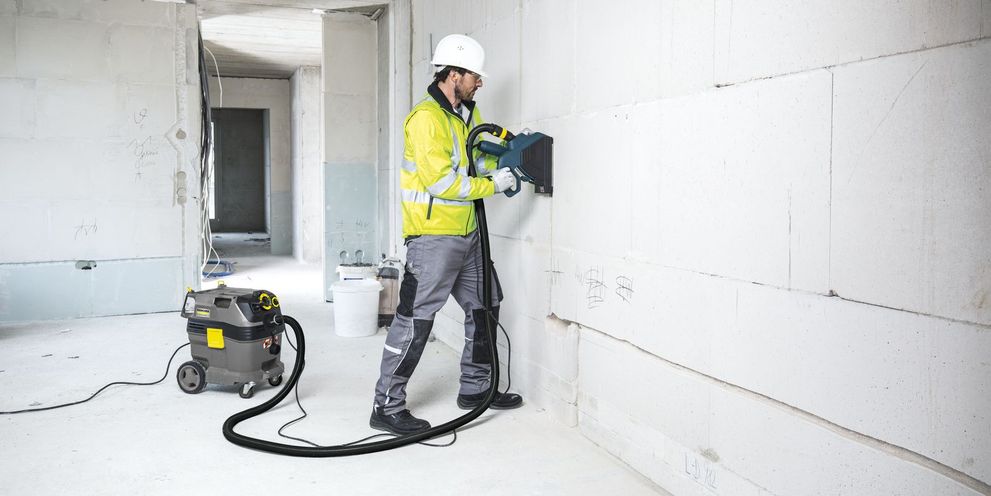 A man in construction uniform cleans a wall with a safety vacuum cleaner