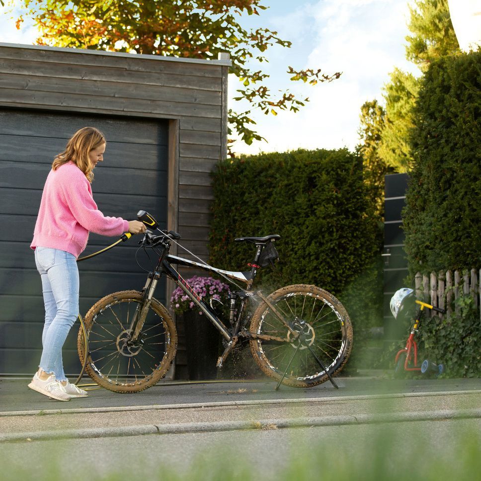 A woman cleans her bike in front of the garage with a Kärcher Mobile Outdoor Cleaner