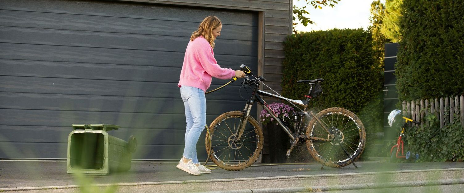 A woman cleans her bike in front of the garage with a Kärcher Mobile Outdoor Cleaner