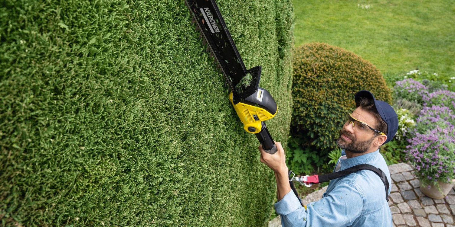 A man works with the Kärcher battery hedge trimmer overhead and uses a shoulder belt