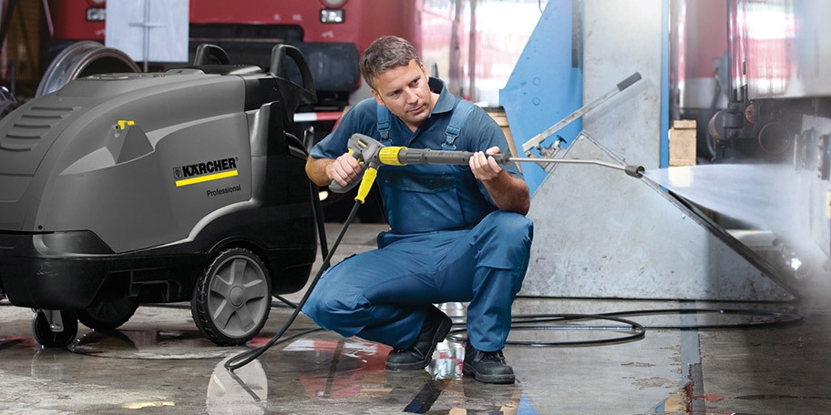 A worker uses a hot water pressure washer to clean the underside of a semi truck.