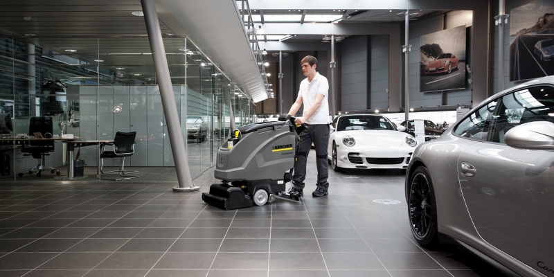 A worker uses a walk-behind floor scrubber to clean inside a showroom.