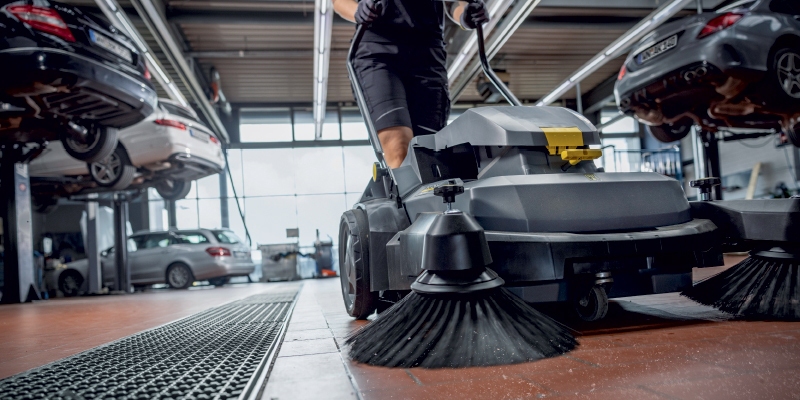A floor-level photo taken of a walk-behind sweeper cleaning inside a service repair shop.